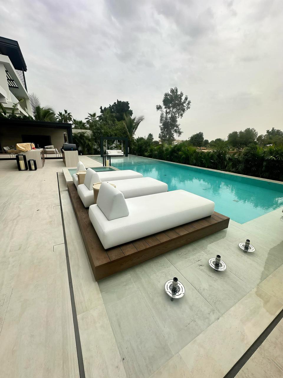 Modern poolside swing with white cushions hanging over clear blue swimming pool surrounded by tropical plants and white lounge chairs.