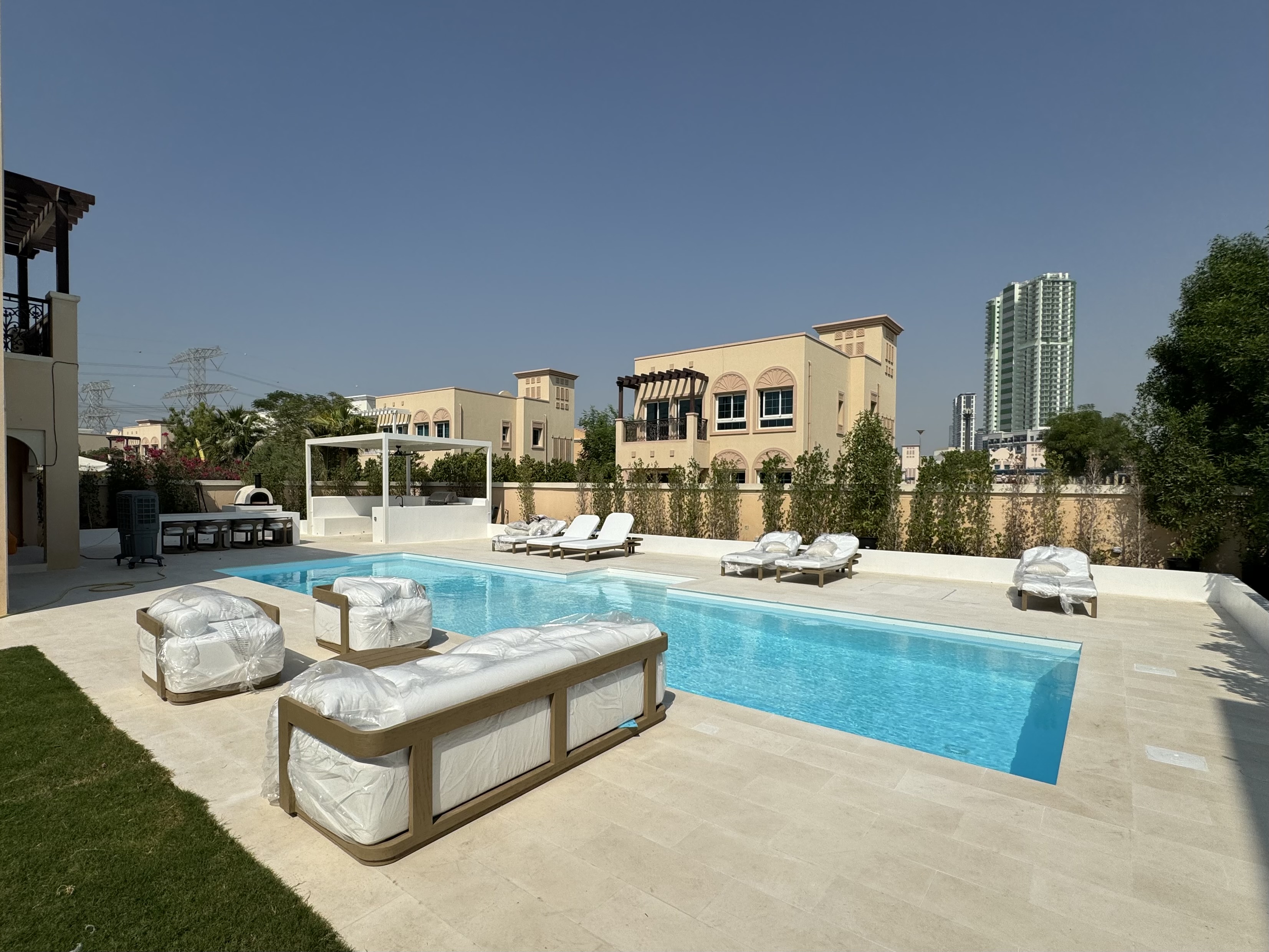 Modern beige villa with multiple balconies beside a clear blue swimming pool and sun loungers wrapped in plastic on a sunny day.