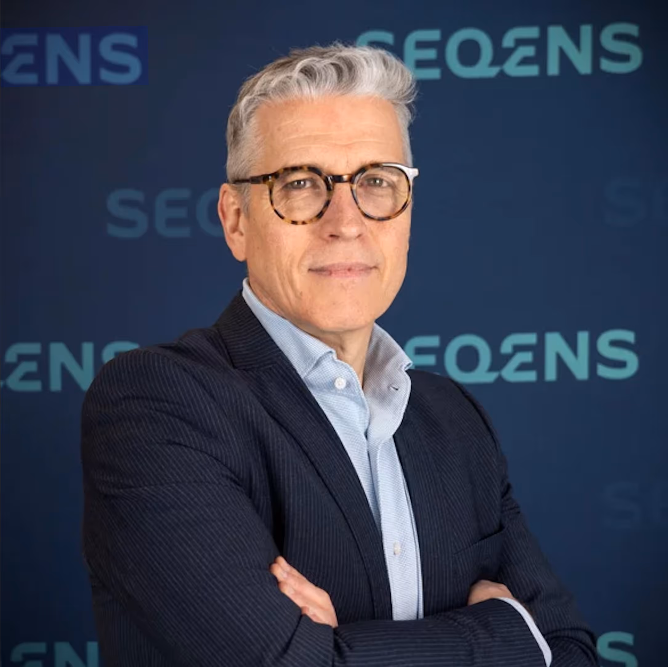 Pascal-Villemagne Homme d'âge moyen aux cheveux gris, portant des lunettes rondes et un costume bleu foncé, posant devant un fond avec le logo SEQENS.