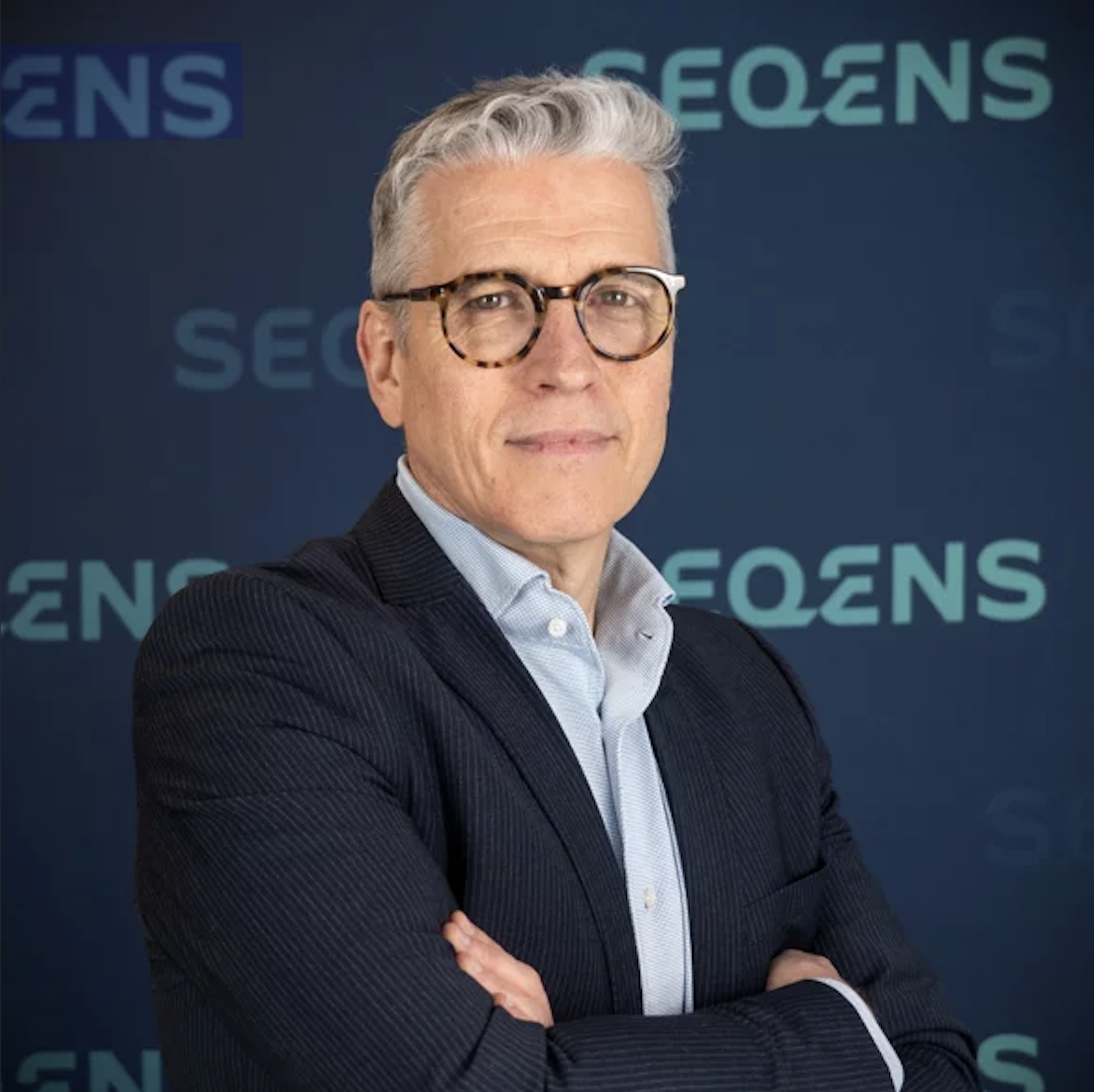 Pascal-Villemagne Homme d'âge moyen aux cheveux gris, portant des lunettes rondes et un costume bleu foncé, posant devant un fond avec le logo SEQENS.