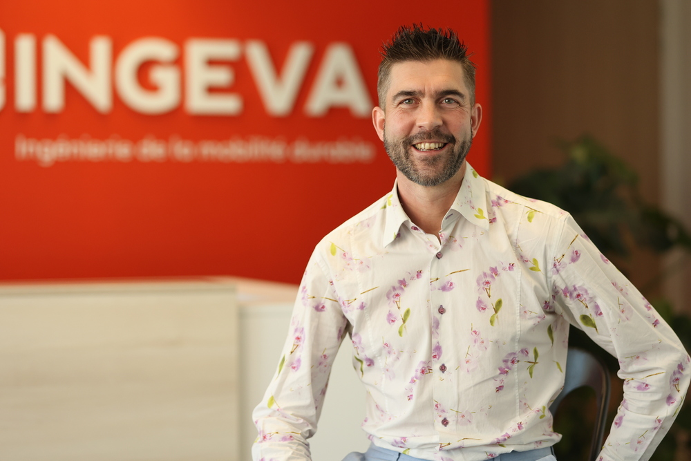 Julien L, Homme souriant en chemise à fleurs assis devant un mur rouge avec le logo INGEVA flou.
