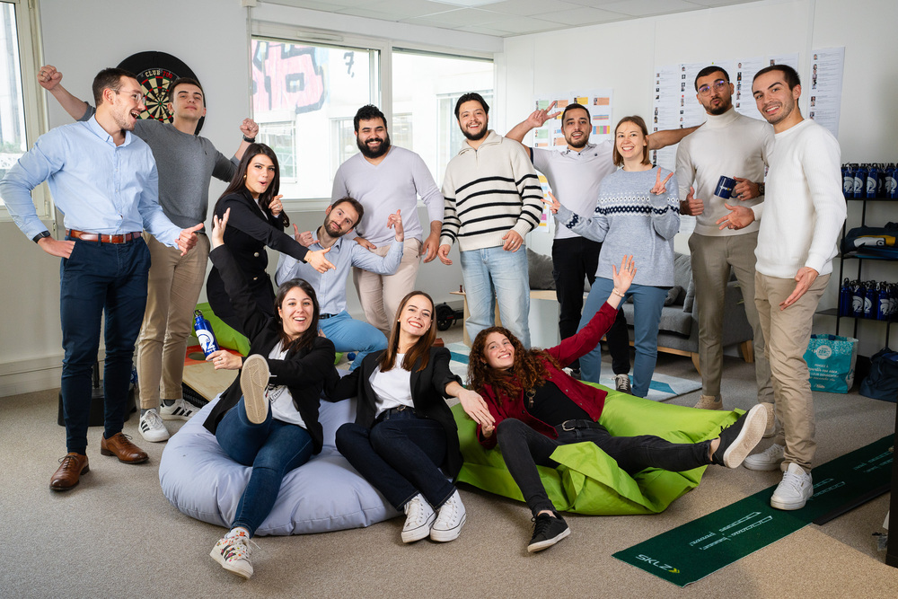 Groupe de jeunes adultes souriants dans un bureau moderne, certains assis sur des poufs, posant de manière conviviale et détendue.