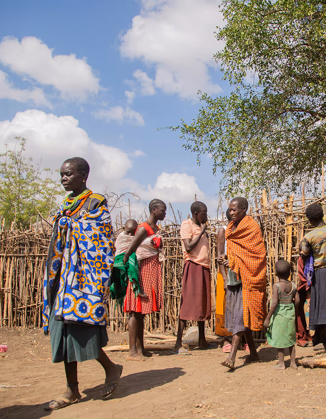 Group of African women and children standing outside near a wooden fence under a partly cloudy sky.