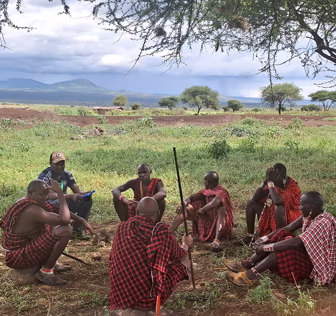 Group of seven Maasai men sitting on the ground in a circle under a tree in a rural landscape with mountains in the background.