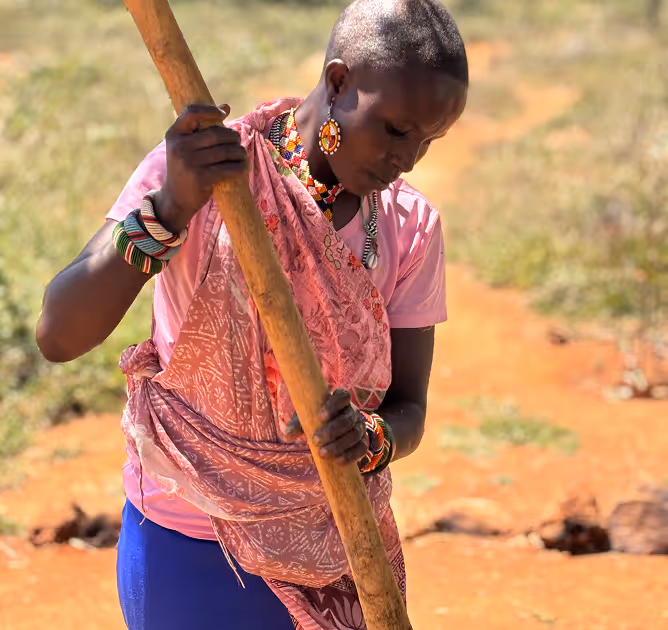 Woman in traditional attire holding a wooden stick outdoors on orange soil.