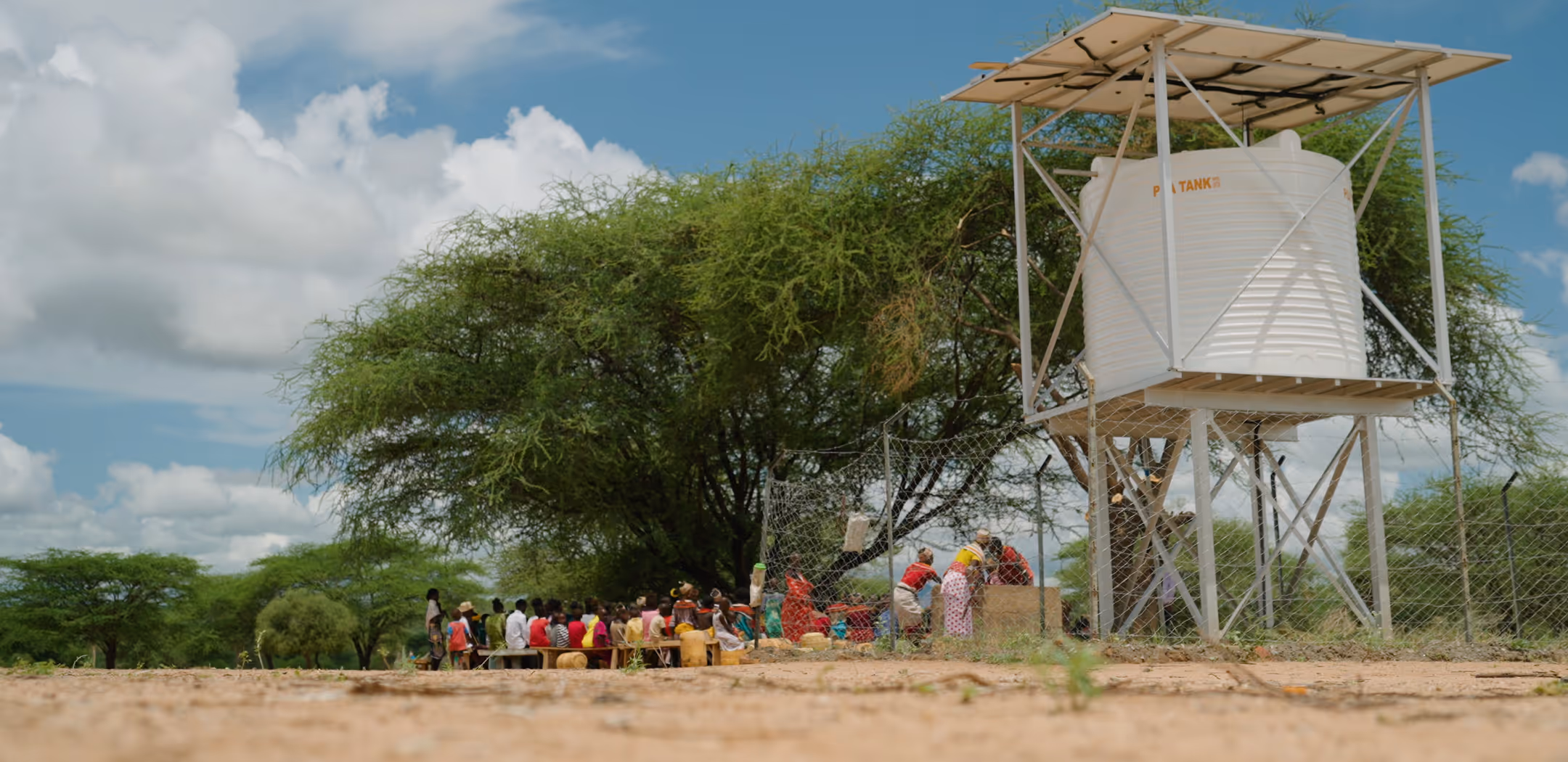 Group of people gathered near a fenced water tank elevated on a metal stand under a large tree in a rural area.