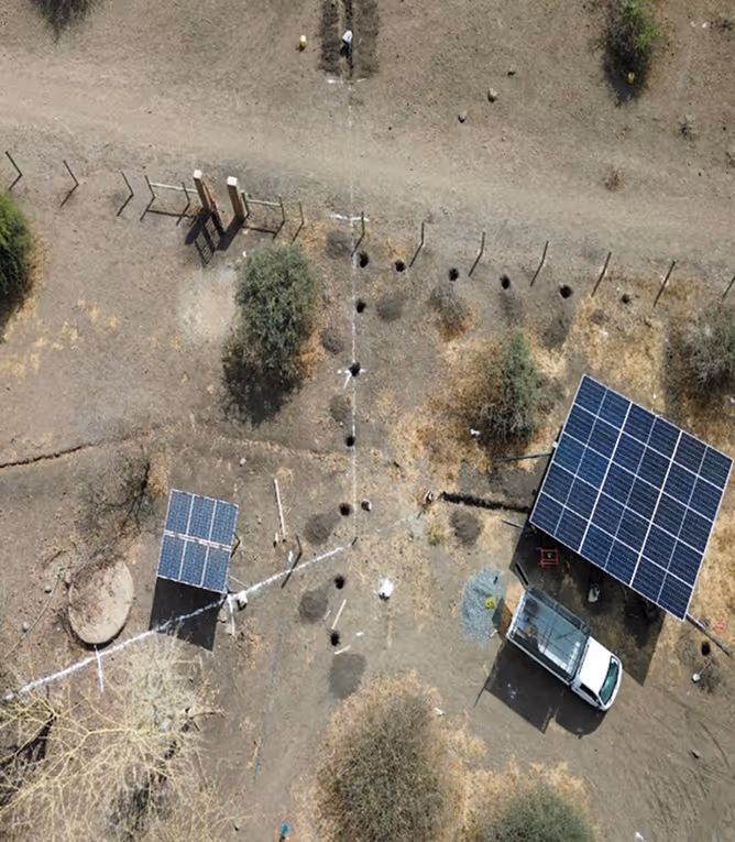 Aerial view of dry terrain with two solar panel installations and a white truck parked nearby, surrounded by scattered holes and sparse trees.