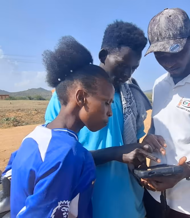 Three people outdoors looking at and interacting with a handheld electronic device, with a rural landscape in the background.