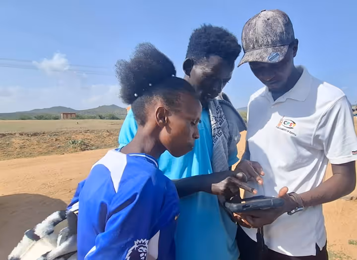 Three young people outdoors looking intently at a handheld electronic device against a backdrop of open land and clear sky.