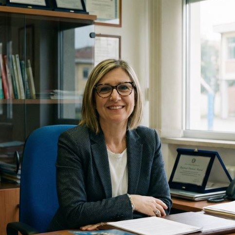 Donna, dirigente scolastico, sorridente con occhiali e giacca seduta a una scrivania con documenti e una targa dietro, in un ufficio luminoso.