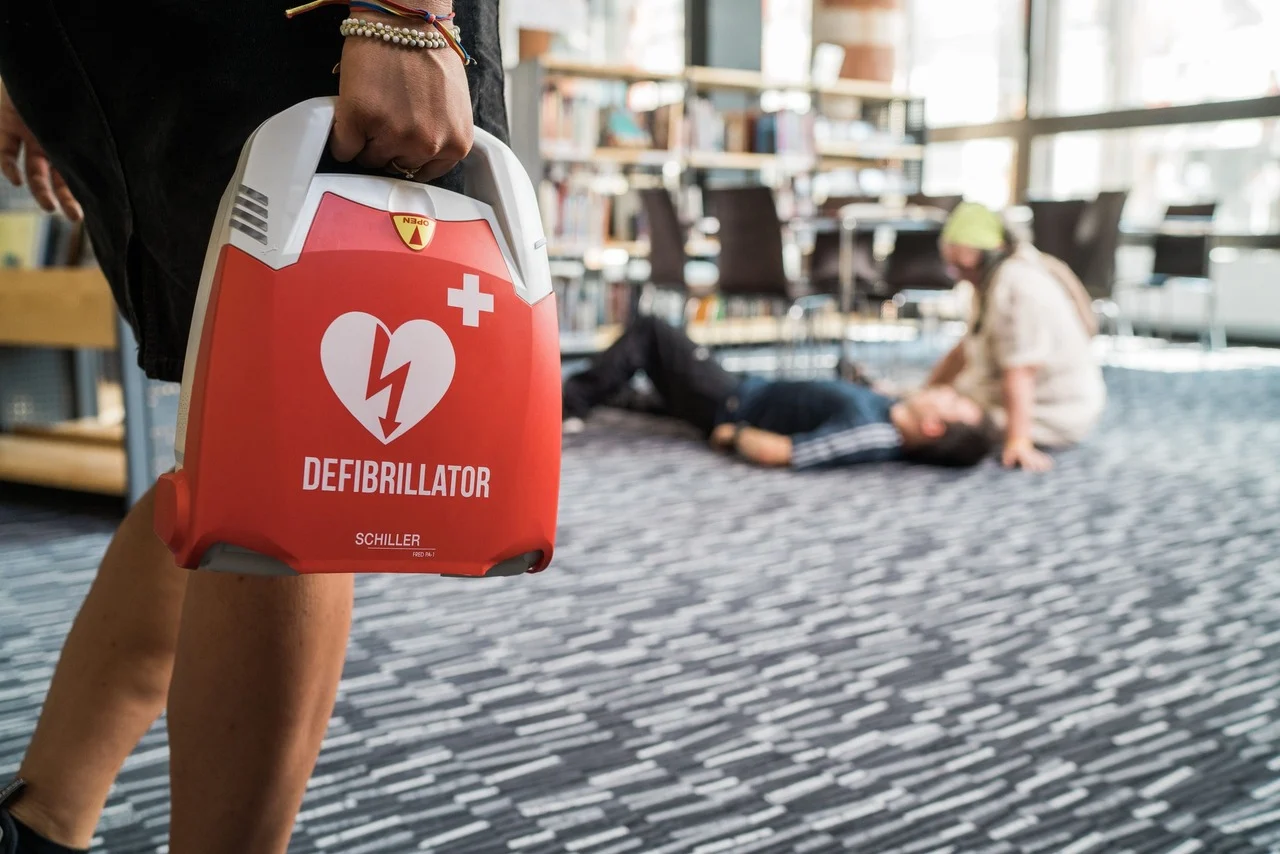 Persona con defibrillatore Schiller FRED PA-1 in mano mentre un'altra persona chiama aiuto vicino a un uomo sdraiato a terra.