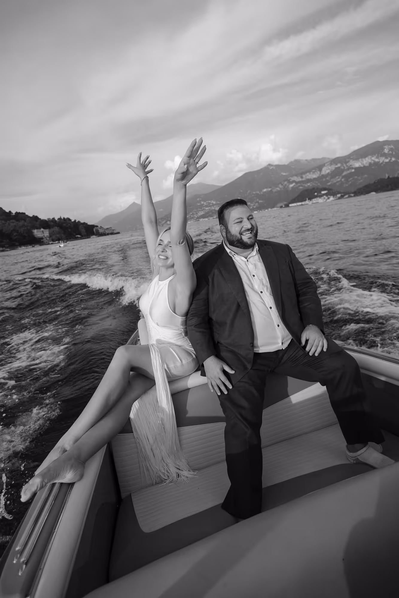 A joyful couple sitting on the back of a boat with water and mountains in the background, the woman raises her arms happily.