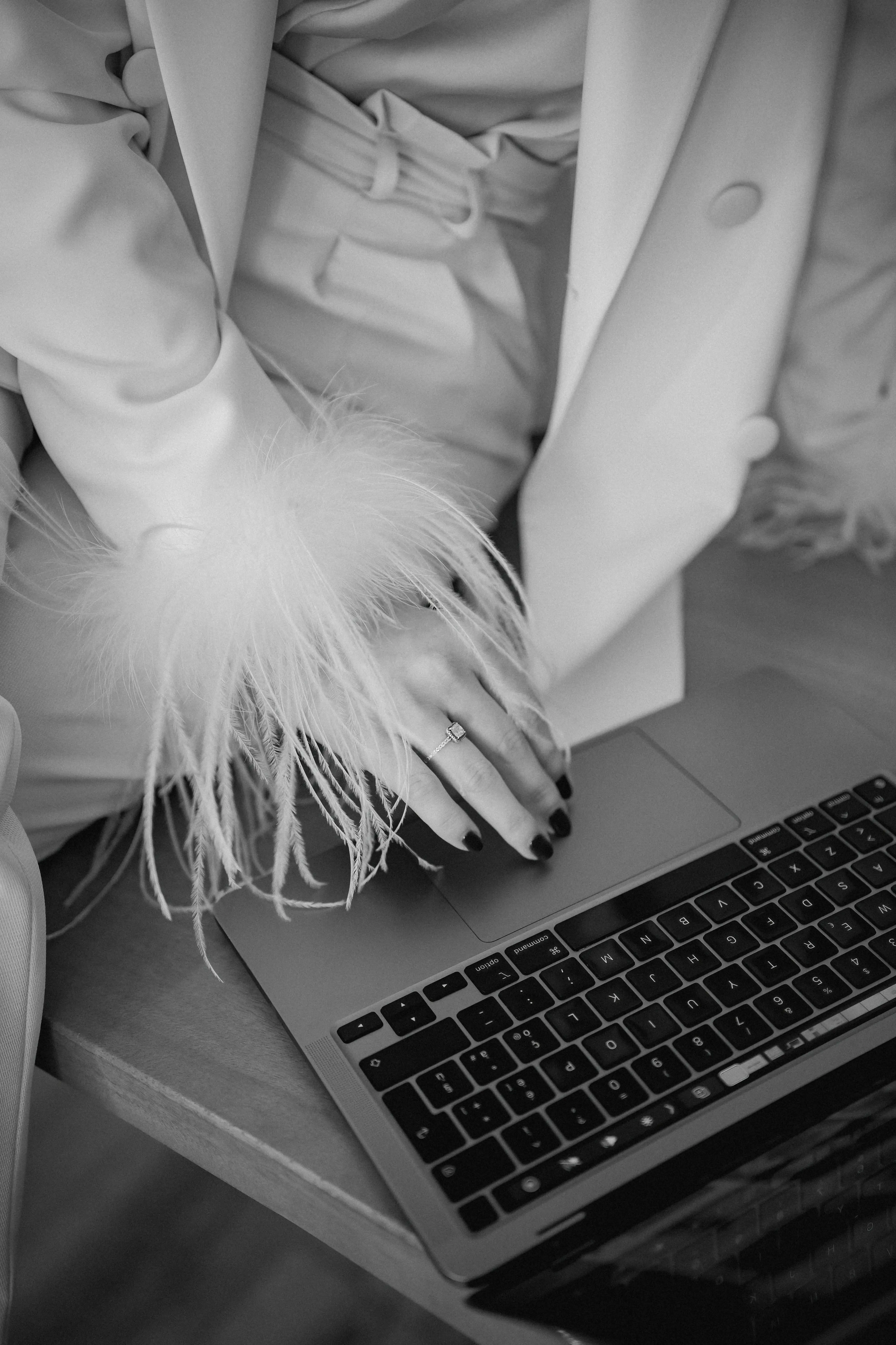Close-up of a hand with black nail polish and a square-cut ring resting on a laptop keyboard, wearing a feather-trimmed sleeve and a belted jacket.