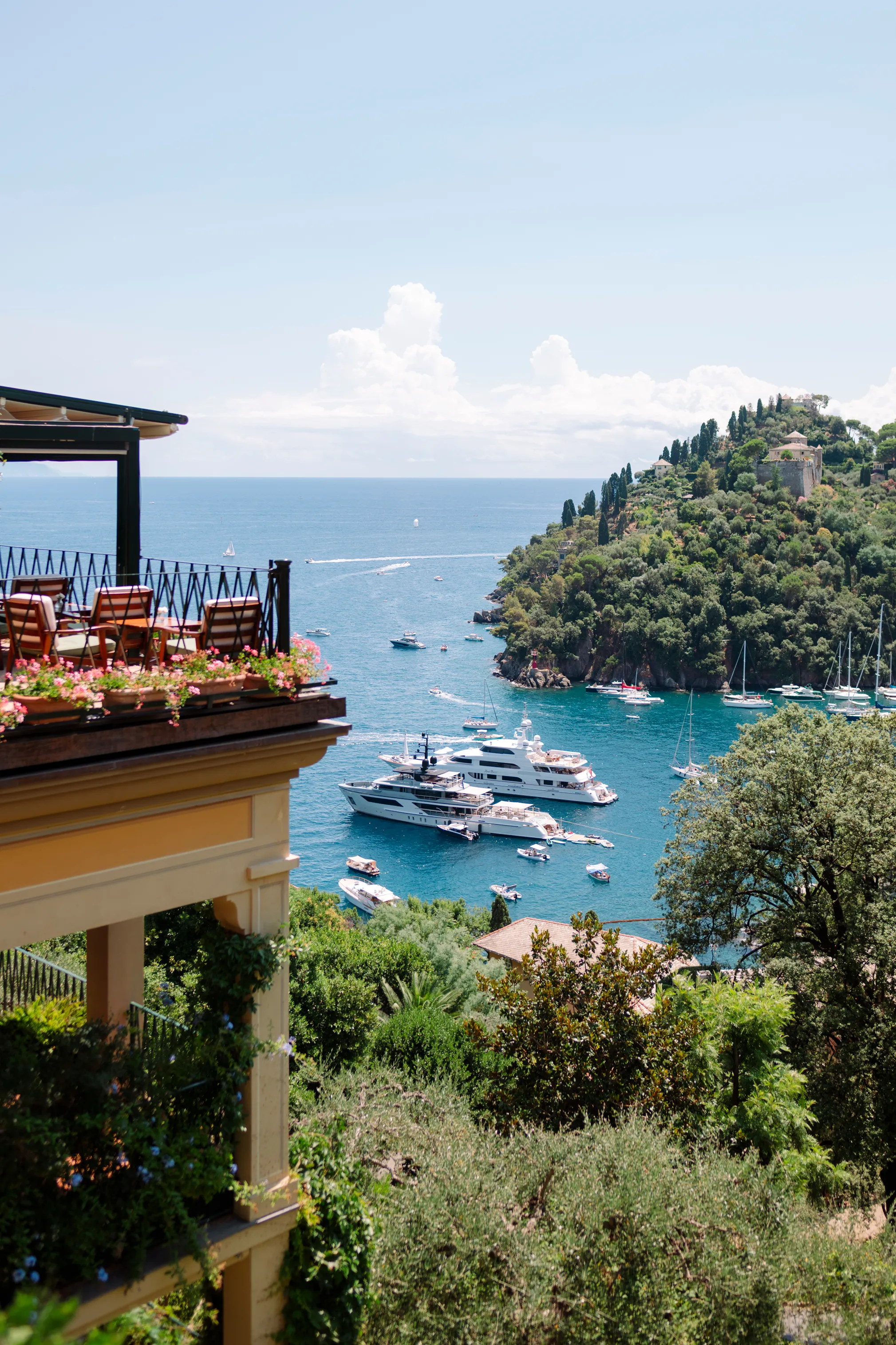 View of a terrace with chairs overlooking a bay filled with yachts and sailboats near a lush green hillside under a clear blue sky.