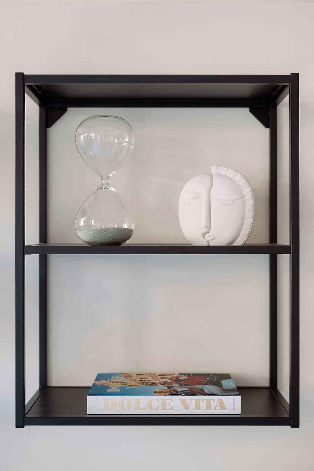 Black metal wall shelf with a clear hourglass and a white ceramic face sculpture on the top shelf, and a book titled 'Dolce Vita' on the bottom shelf.
