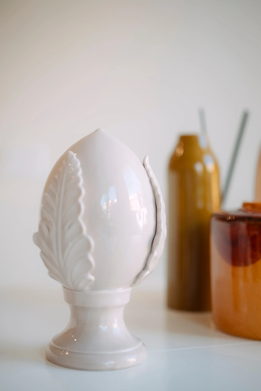 White ceramic decorative object shaped like a stylized artichoke with leaf motifs, placed on a white surface with blurred brown and amber glassware in the background.