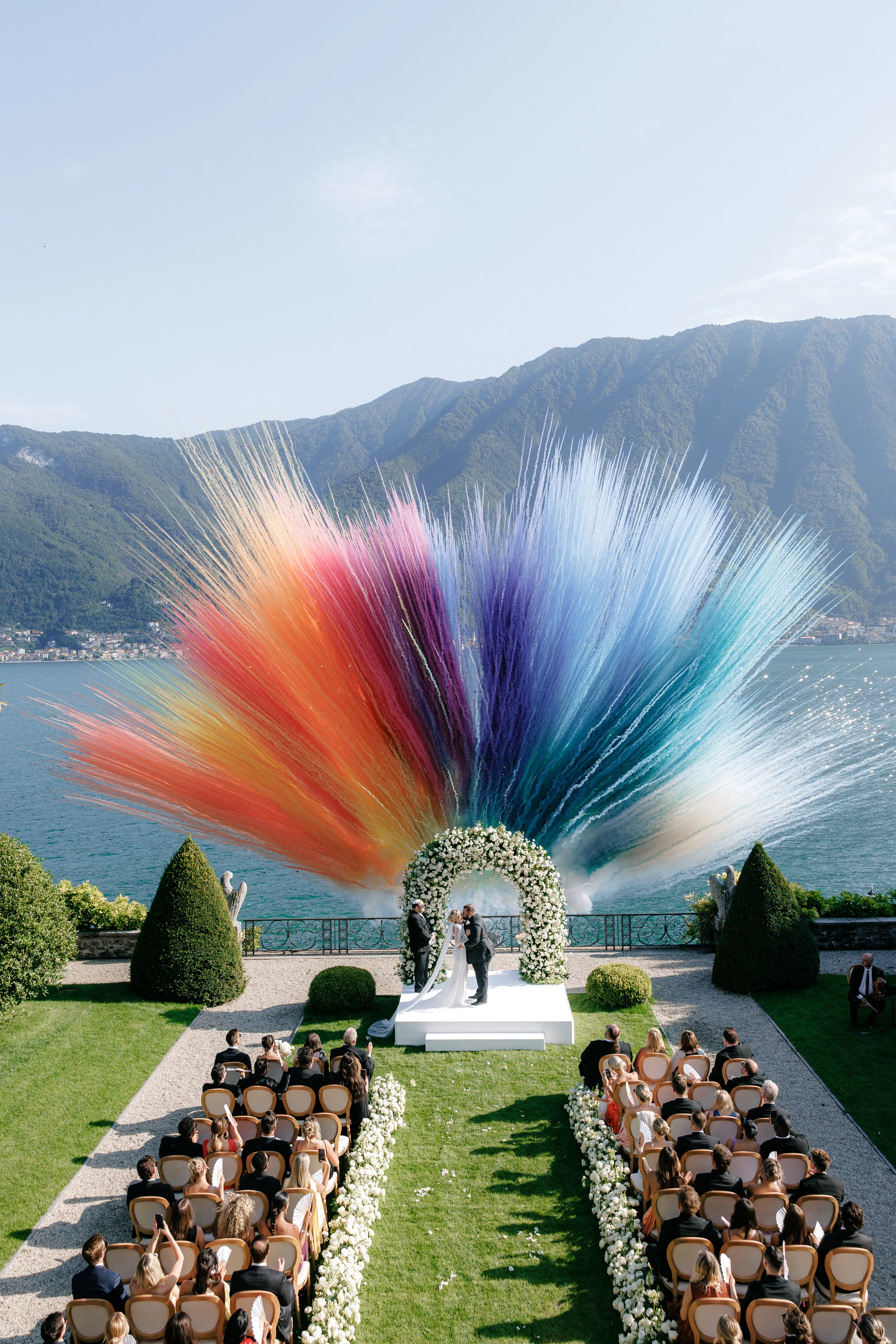 Outdoor wedding ceremony by a lake with mountains in the background and colorful fireworks bursting behind the couple under a white floral arch.