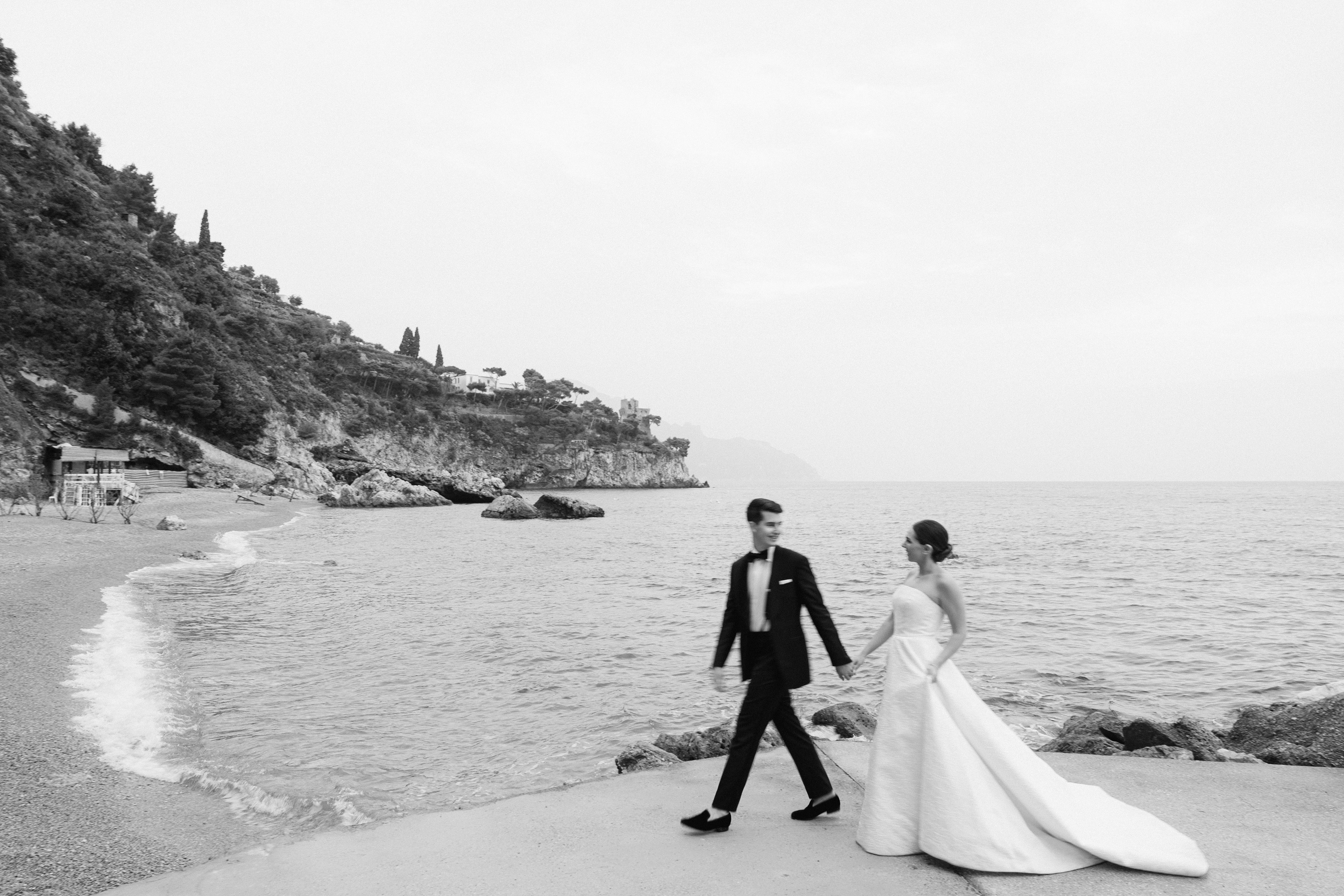 Newlyweds walking hand in hand on rocky coastal beach in Amalfi