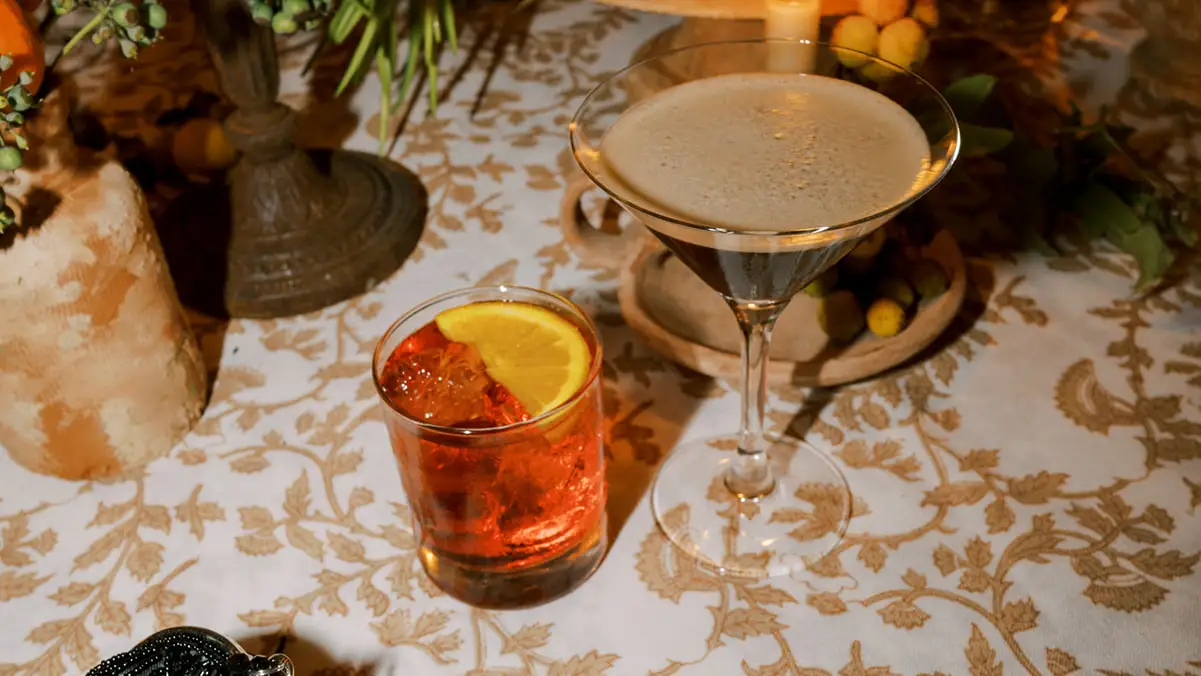Two cocktails on floral tablecloth: one orange, one frothy martini glass