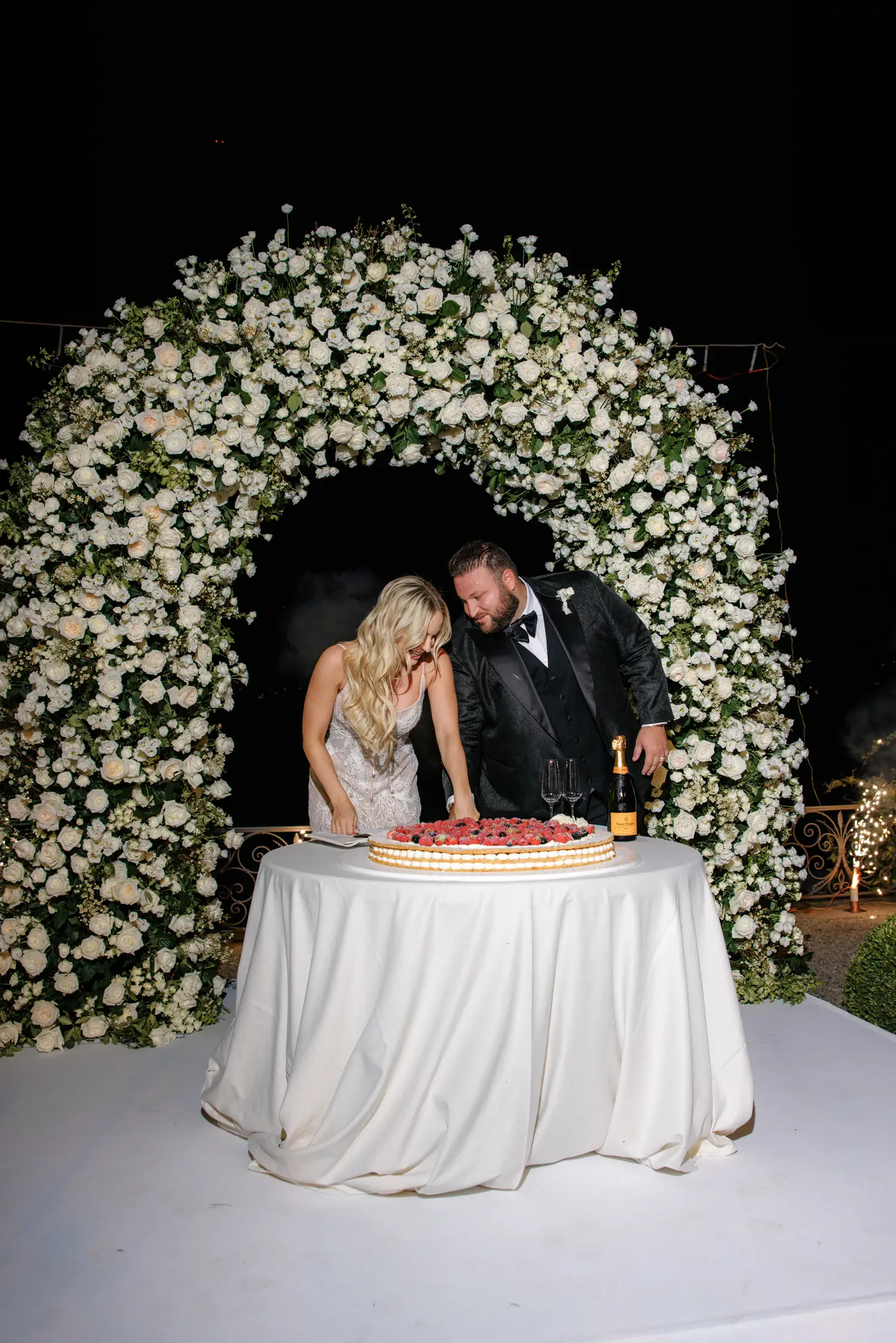 Wedding couple cutting cake under floral arch at elegant night ceremony on Como Lake