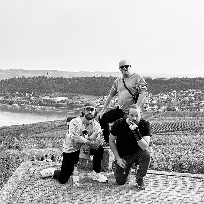 Three Artus employees posing on a paved overlook with a river, vineyard rows, and town in the background.