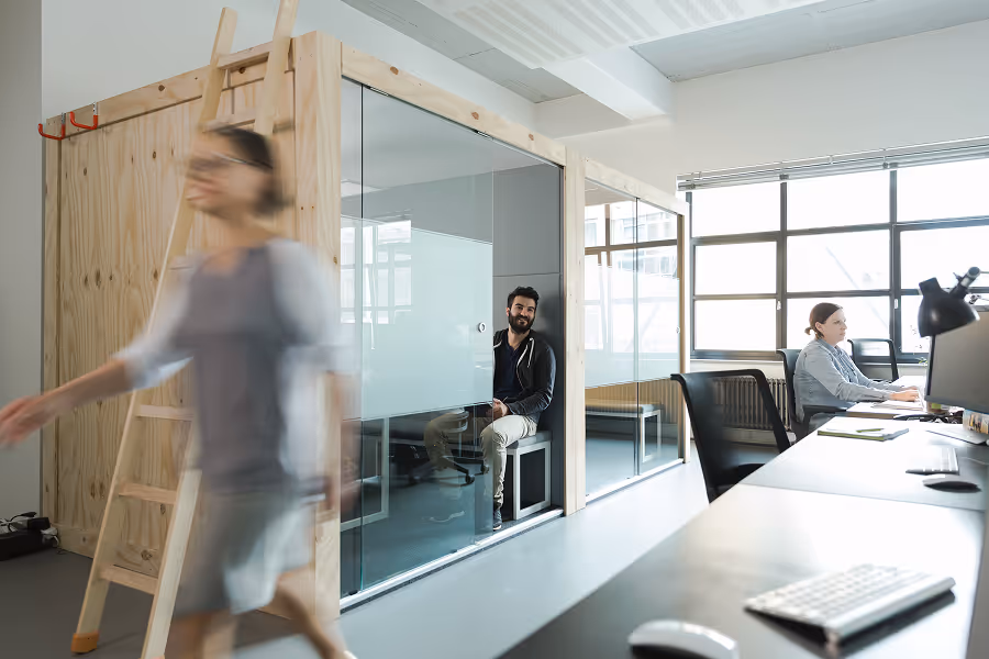 Modern office with people working at desks, one man sitting in glass-walled meeting room, woman walking by.
