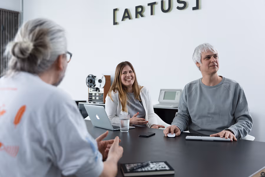 Three people in a meeting room, one woman and two men talking around a table with a laptop and keyboard.