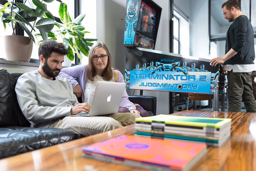 Two people sitting on a couch working on a laptop, a third stands playing a Terminator 2 pinball machine.