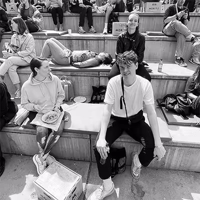 Group of young Artus employees sitting and relaxing on outdoor wooden steps, with some eating and chatting.