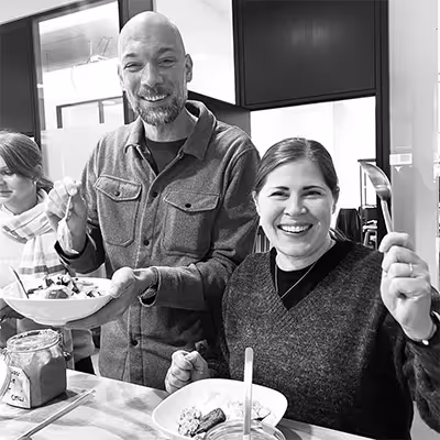 Two smiling Artus employees holding forks and plates of food while sitting at a table in a kitchen.
