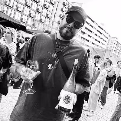 Artus employee wearing sunglasses and a cap holds a wine glass and a bottle at an outdoor event with people in the background.