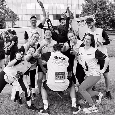 Group of nine happy Artus employees in matching event shirts posing outdoors on grass, some holding drinks and making peace signs.