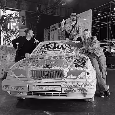 Three Artus employees posing with a white car covered in graffiti inside an industrial or event space; one person leans on the car, another sits on the hood making a peace sign, and the third stands to the side.