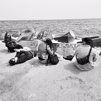 Four Artus employees sitting and lying on a sandy beach in Barcelona near graffiti-covered rocks by the sea.