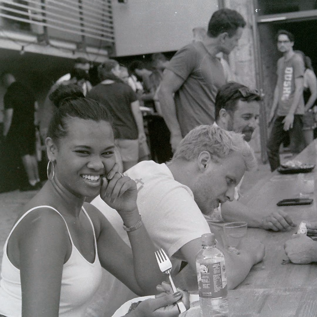 Smiling woman with a fork sitting at a crowded outdoor table with others in black and white photo.
