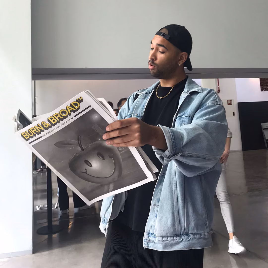 Man in a denim jacket and black cap reading a newspaper titled Burn & Broad with a smiling apple image.