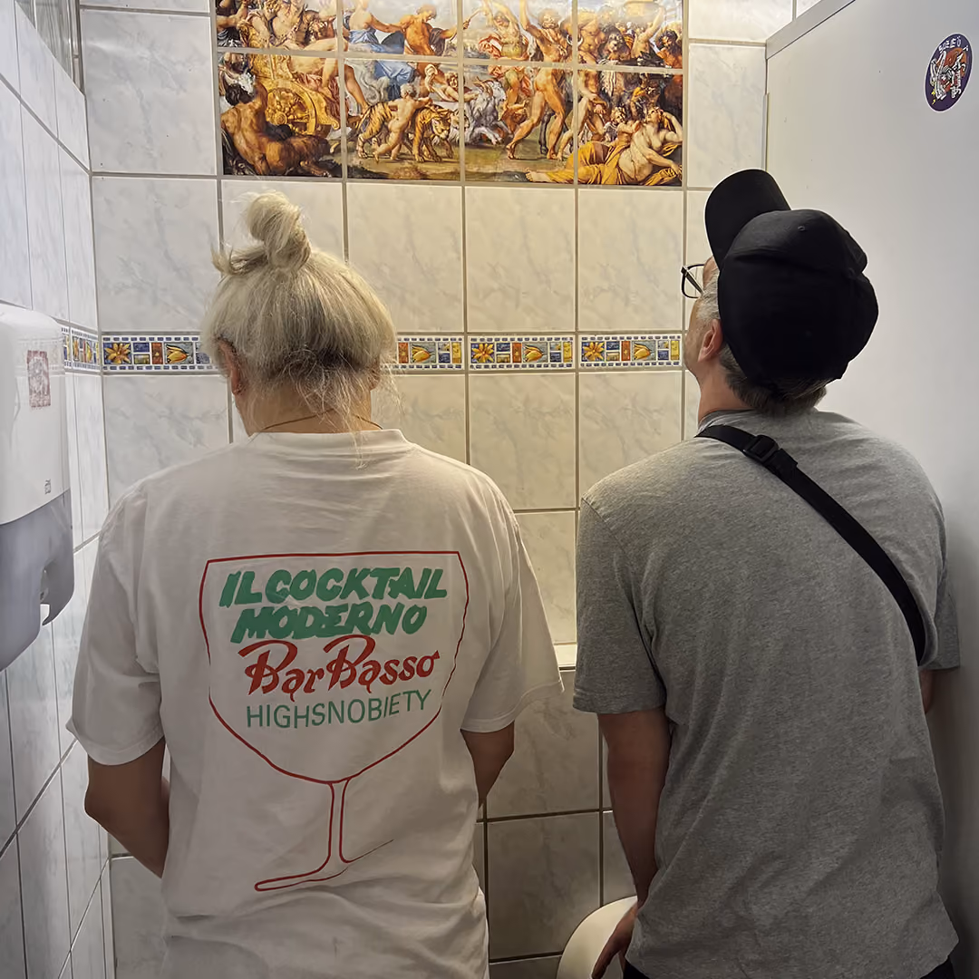 Two people standing side by side in a tiled restroom urinal area, one wearing a white graphic tee.