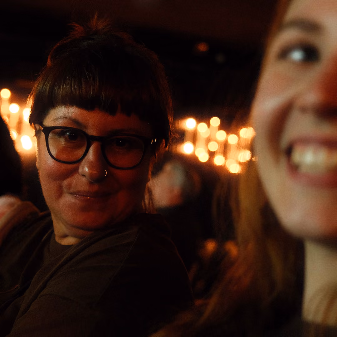 Two women smiling in dim light with blurred string lights in the background.