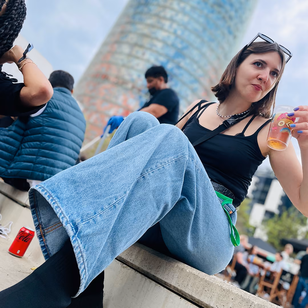 Young woman in black top and wide jeans sitting outside, holding a drink with a colorful tower in background.
