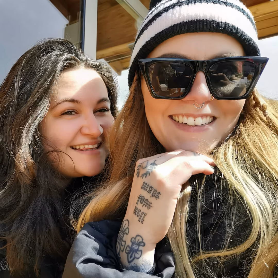 Two smiling women, one with dark hair, the other with sunglasses, beanie, and visible hand tattoos.