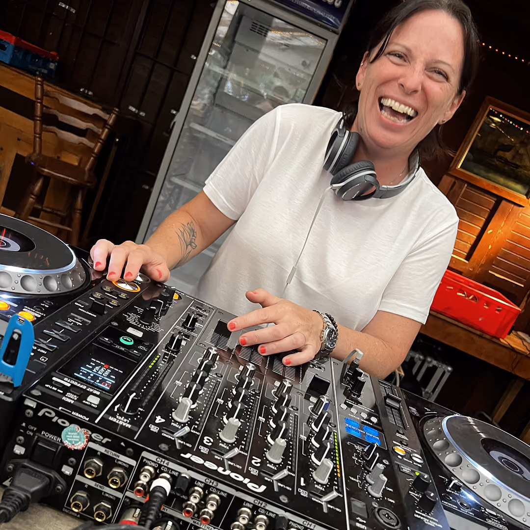 Woman smiling while using DJ mixer with headphones around her neck and red nail polish.