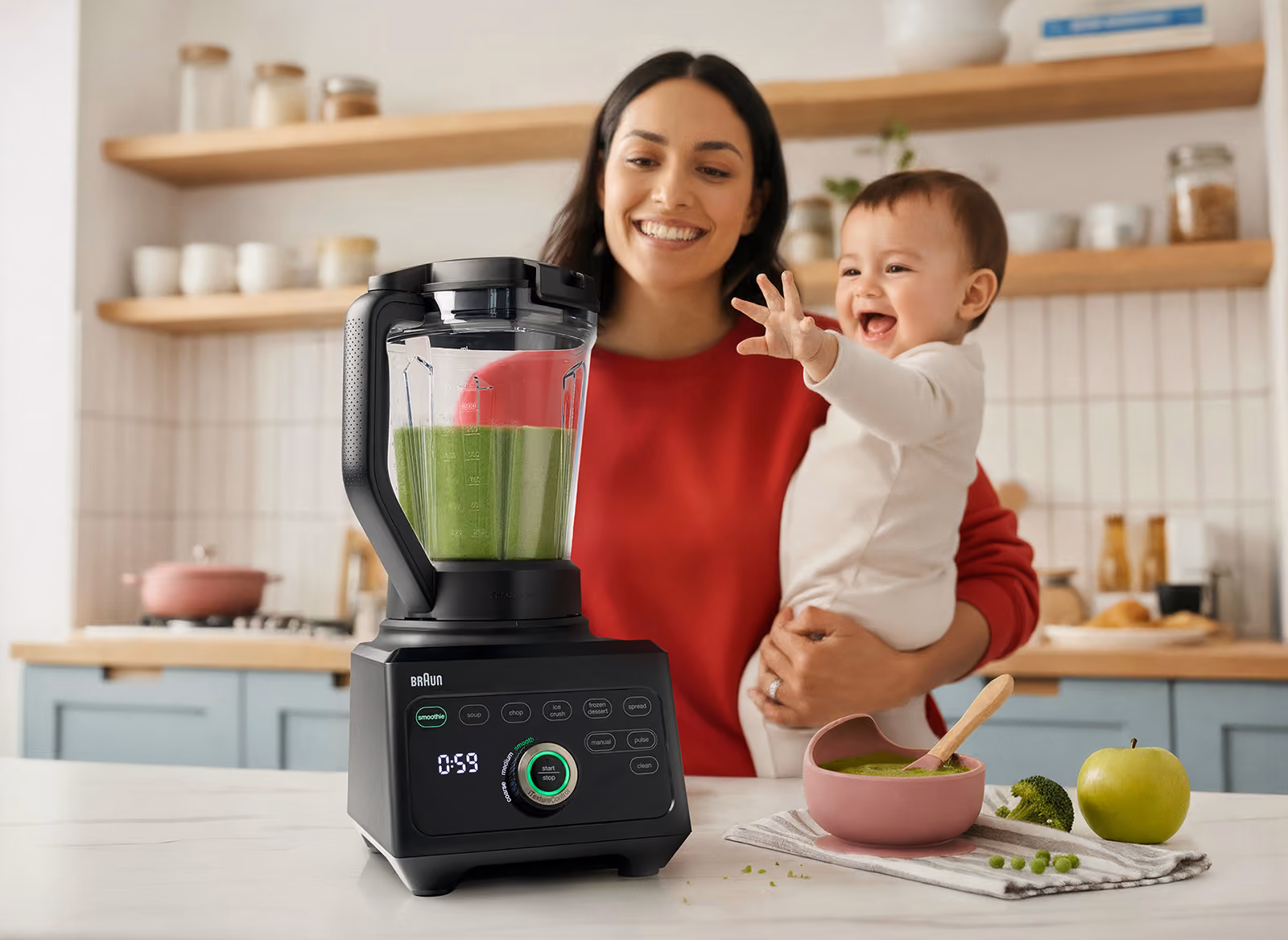 Smiling woman holding baby next to blender with green smoothie and bowl of baby food on counter.