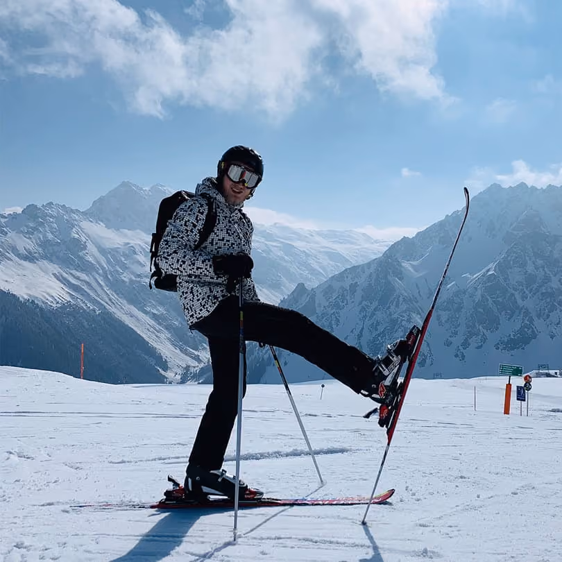 Skier in patterned jacket on snow-covered mountain slope lifting one ski in front against clear sky.