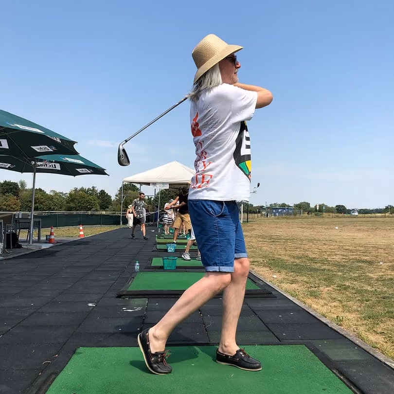 Person in hat and shorts swinging a golf club at a driving range on a sunny day.