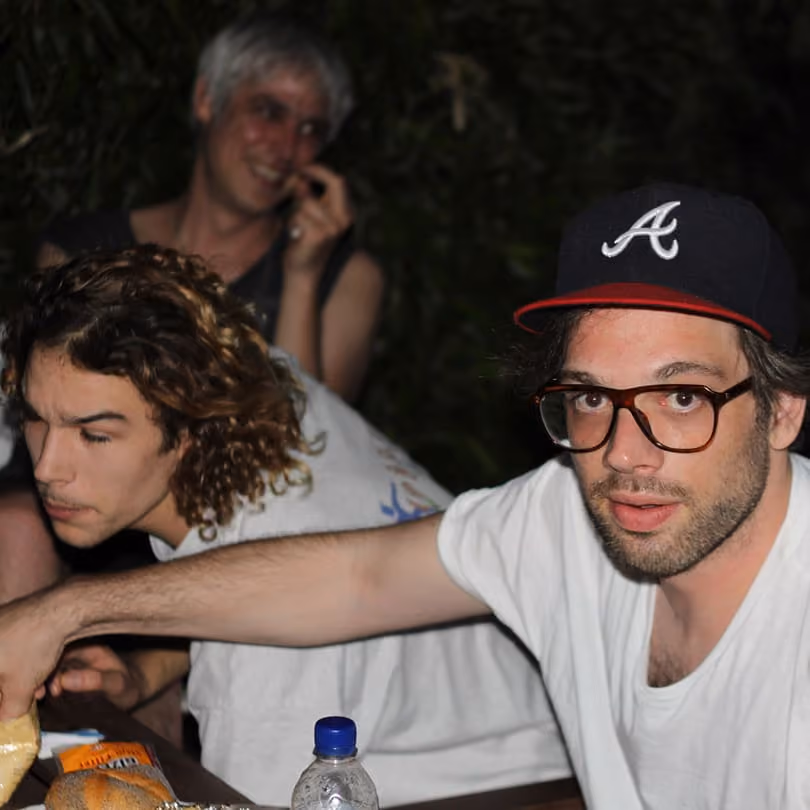 Three people sitting at a table with food, one wearing glasses and a dark baseball cap with an A.
