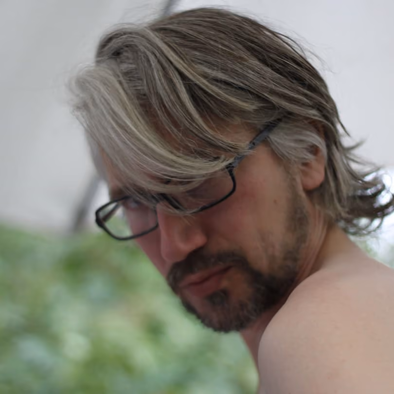 Man with light brown hair, beard, and glasses looking over his shoulder outdoors.