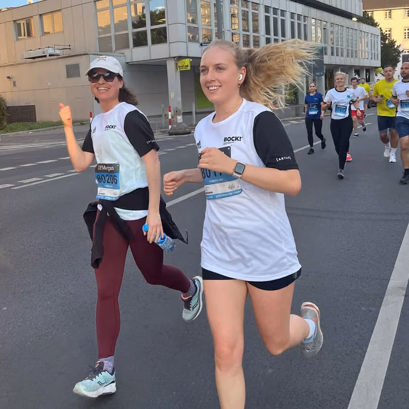 Two women smiling and running on a city street during a race with other runners behind them.