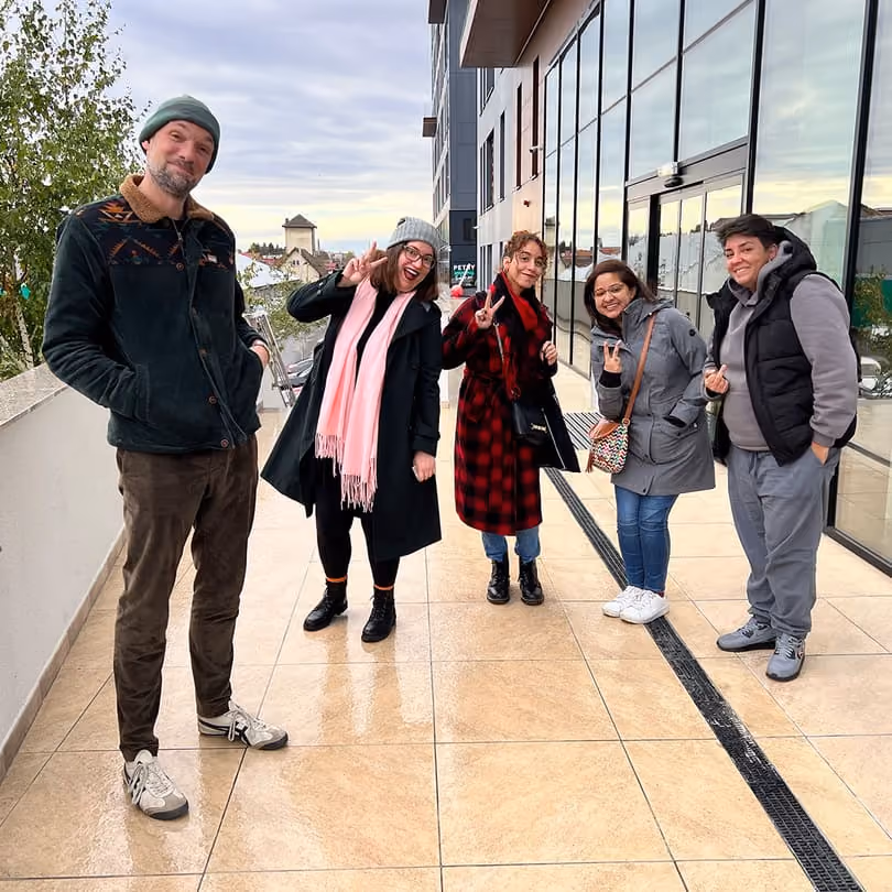 Five people outdoors on tiled patio, smiling and making peace signs near glass building.