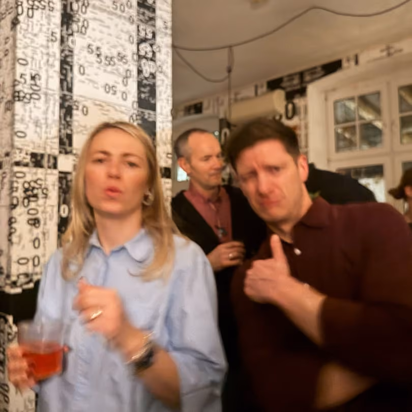 Two people at a party, one giving a thumbs up and another holding a drink, blurred motion.