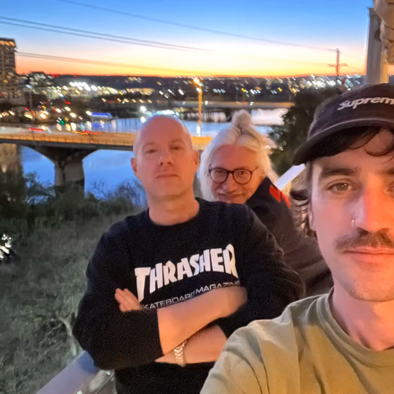 Three people posing on a balcony at sunset with a city and bridge over water in the background.